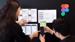 Dos diseñadores colaboran revisando wireframes impresos sobre una pared negra, discutiendo esquemas de una app móvil con colores y estructuras. El logo de Figma aparece en la esquina superior derecha.