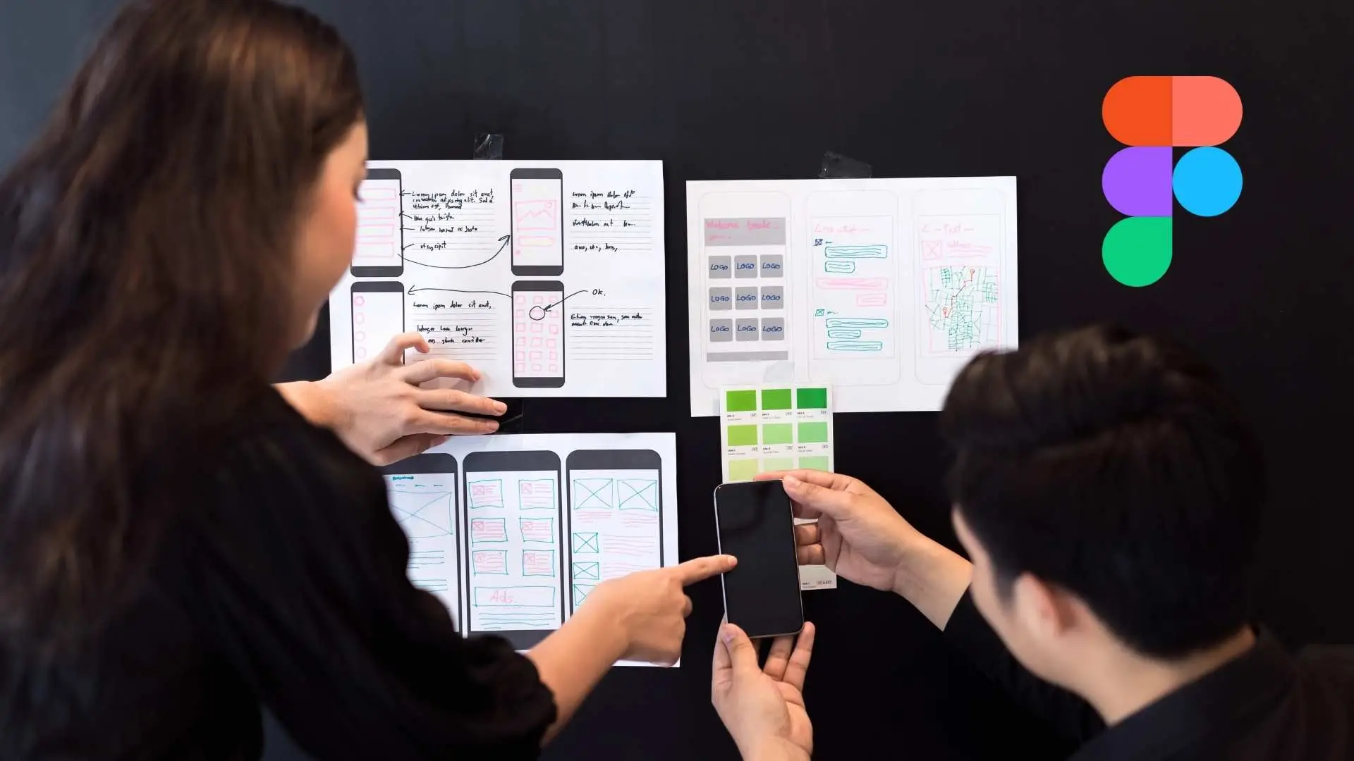 Dos diseñadores colaboran revisando wireframes impresos sobre una pared negra, discutiendo esquemas de una app móvil con colores y estructuras. El logo de Figma aparece en la esquina superior derecha.